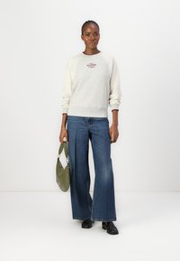 Sweat-shirt gris clair avec des manches crème, arborant un logo rouge. Associé à un jean bleu à jambes larges et un sac à main vert. Des chaussures noires complètent le look.