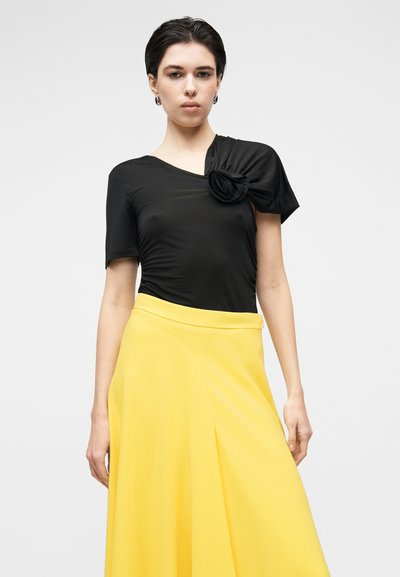 Haut noir à manches courtes avec un accent floral drapé sur l'épaule, associé à une jupe jaune fluide à coupe légèrement trapèze.