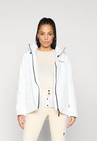 Mulher com cabelo trançado, vestindo uma jaqueta branca com capuz sobre uma blusa de chiffon creme e calças bege claras com botões pretos e logotipo.