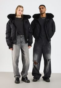 To unge voksne i sorte jakker med pelskapper, sorte toppe og løse, falmede jeans står side om side på en neutral baggrund.