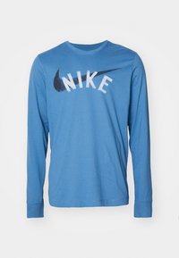 Modrá dlouhá rukávová košile Nike s bílým textem Nike a černým logem swoosh přes hruď, umístěná na bílém pozadí.