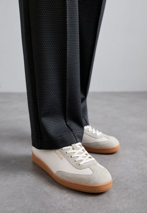 Hvide og grå sneakers med suede-detaljer og en gummisål, parret med mørke mønstrede bukser. Skoene har snøre-design og logo-detajle.