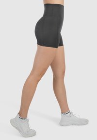 Pantaloni sportivi neri a vita alta con una texture liscia e un design aderente, abbinati a sneaker bianche con un dettaglio in nero.