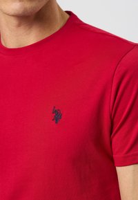 Close-up of person wearing a red t-shirt with a small embroidered dark logo of a polo player holding a mallet on the chest.