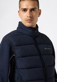 Mężczyzna w granatowej pikowanej kamizelce Champion zapinanej na zamek, noszący granatową koszulę z długim rękawem i białymi akcentami, o neutralnym wyrazie twarzy, na białym tle.