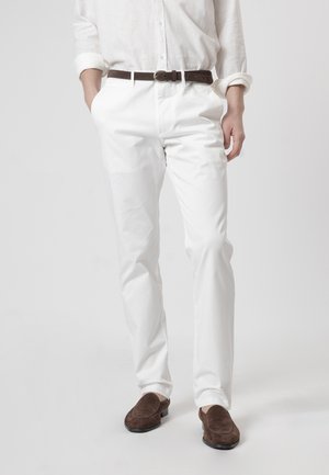 Hombre de pie con las manos en los bolsillos, lleva pantalones blancos, camisa blanca abotonada con las mangas remangadas, cinturón marrón y mocasines marrones.