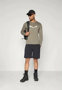 Long-sleeve olive green shirt with a light logo, paired with black shorts, black boots, and a black cap. Model carries a backpack.