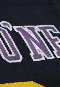 Schwarzer Stoff mit großen, weißen und lila gestickten Buchstaben "UNF", darunter ein gelber Akzent mit glatter Textur.