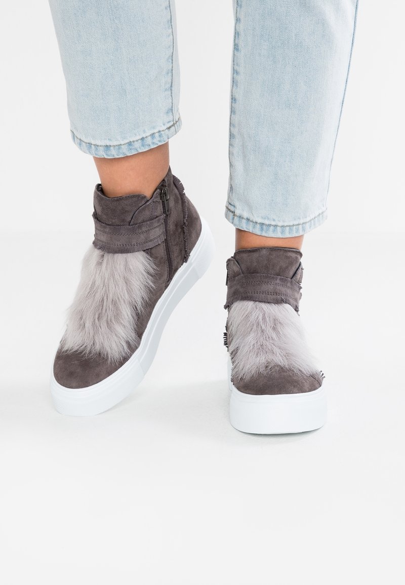 Chaussures à chevilles en daim gris avec des accents en fausse fourrure, semelle en caoutchouc blanche et fermeture éclair sur le côté. Jeans retroussés au-dessus des chaussures.