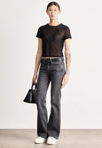 Čierny pruhovaný sieťovaný crop top, sivé svetlé džínsové nohavice s vysokým pásom a rozšíreným strihom, čierny opasok, biele tenisky, drží malú čiernu kabelku. Hladké textúry.