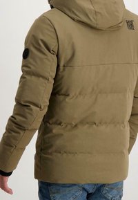 Tajtat passande olivgrön puffjacka med huva, rutigt mönster i sömmarna, svart logotyppatch på ärmen och ribbade ärmslut.