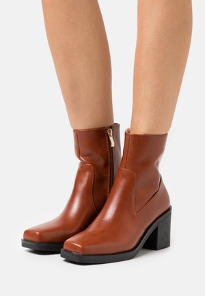 Bottes courtes en cuir marron avec un bout carré, une fermeture éclair latérale et un talon noir épais. Texture lisse et design minimaliste.