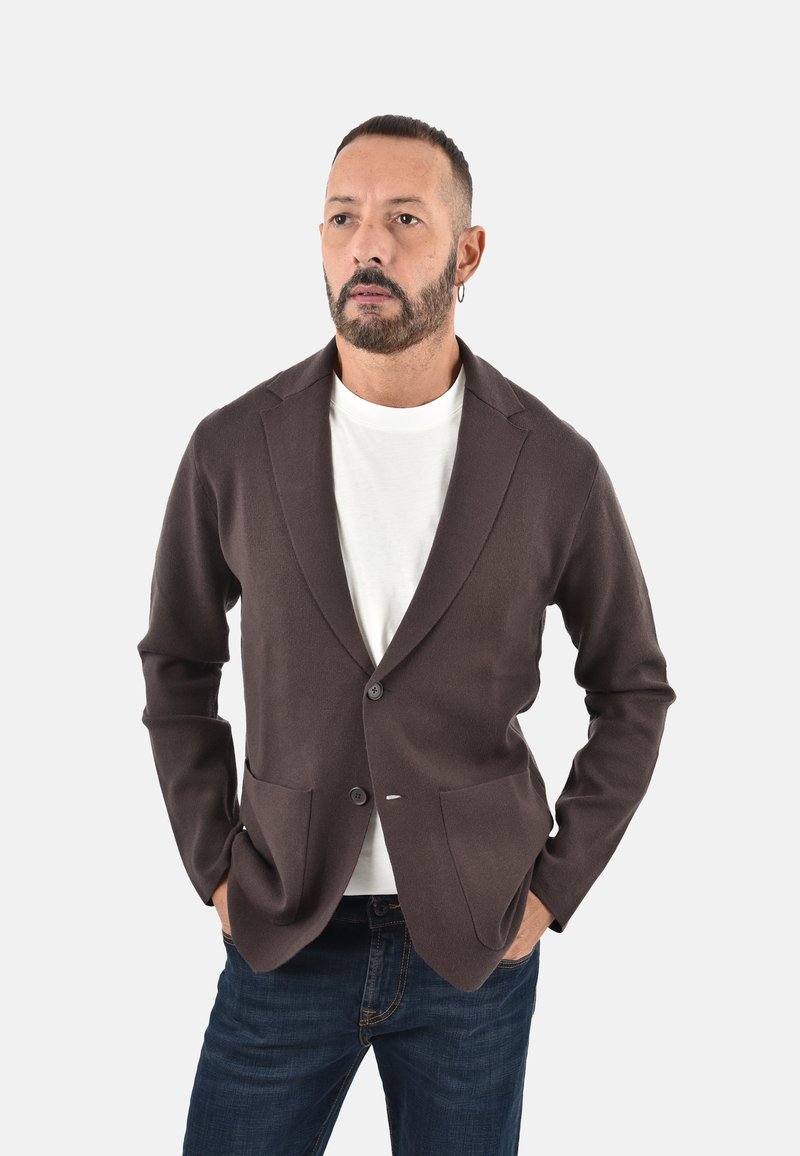 Blazer marrone in maglia con vestibilità su misura, rever con intaglio, due tasche frontali e chiusura con bottoni, indossato sopra una camicia bianca e jeans blu.