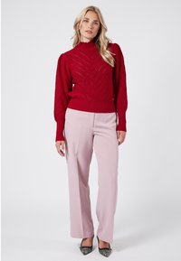 Maglione rosso lavorato a maglia con collo alto, motivo a zigzag e polsini a coste, abbinato a pantaloni palazzo rosa chiaro e décolletés neri a punta.