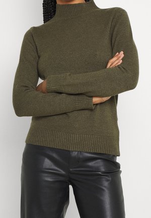 Person med fletninger iført en olivengrøn ribbet sweater og sorte læderbukser, med armene krydsede ved livet mod en ensfarvet baggrund.