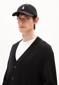 Schwarzer Cardigan mit V-Ausschnitt und Knopfverschluss, mit einem dezenten gestickten Logo auf der Brust. Getragen über einem weißen T-Shirt. Schwarze Kappe auf dem Kopf.