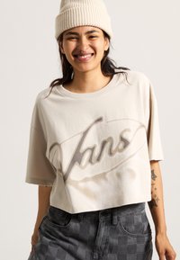 Beige cropped t-shirt with a faded black "Vans" logo, featuring short sleeves and a round neckline, paired with gray checkered pants.