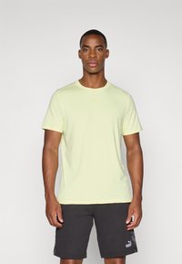 Lys gul sport t-shirt med korte ærmer, rund udskæring og et lille, tonalt logo. Bæres med mørke shorts i ensartet tekstur.
