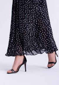 Svart klänning med prickigt mönster i genomskinlig, flödande tyg, med en vågig nederdel. Bärs med svarta lackläder högklackade sandaler.