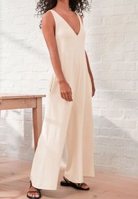 Beige mouwloze jumpsuit met een diepe V-hals, voorzien van brede pijpen en zijzakken. Gekleed met zwarte sandalen, voor een licht bakstenen muur.