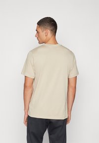 Beige puuvillane T-särk lühikeste varrukatega ja ümmarguse kaelusega, millel on lihtne disain ilma logo või mustriteta, nähtuna tagant.