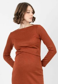 Robe à manches longues en tissu couleur rouille, présentant une silhouette ajustée et un design à l'avant torsadé. Texture douce et décolleté bateau.