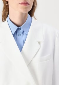 Biely blazer s textúrovaným povrchom, s klopovými lícami a jednohrudníkovou vreckom na hrudi. Pod ním svetlomodrá košeľa s golierom a detailmi na gombíky.
