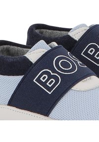 Chaussures de sport en mesh bleu avec une sangle élastique navy arborant le texte "BOS" en blanc. Surface texturée avec des accents crème et zone de renforcement au niveau des orteils.