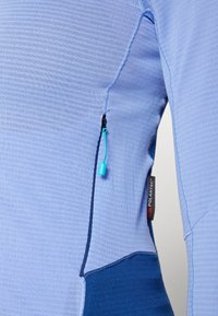 Material textil de culoare albastru deschis cu un buzunar cu fermoar lateral, accentuat de o secțiune de culoare albastră contrastantă și un eticheta Polartec.