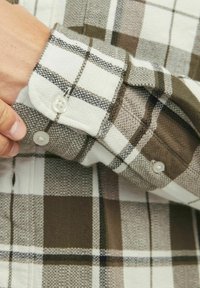 Cămașă cu mânecă lungă, în carouri, cu un model alb, maro și gri. Are manșete cu buton cu butoane albe și o țesătură texturată.