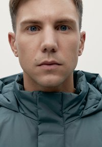 Giacca imbottita grigia con colletto alto e cuciture visibili. La texture è liscia e il design è funzionale senza decorazioni.