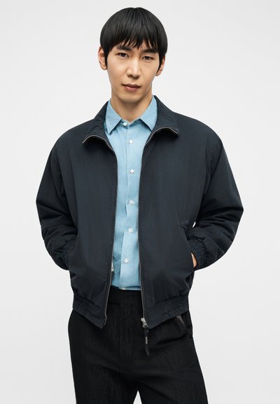 Νέος άνδρας που φοράει μαύρο μπουφάν με φερμουάρ πάνω από ένα ανοιχτό μπλε πουκάμισο και μαύρο παντελόνι, στέκεται με τα χέρια στις τσέπες.