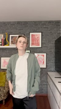 Casaco bomber verde, t-shirt branca, calças pretas. Sala com parede de tijolo escuro, arte emoldurada e uma cadeira de braço com padrão ao fundo.