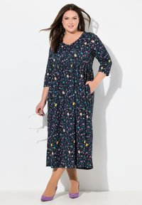 Blumendruck, marineblaue Kleid mit Dreiviertelärmel, geraffter Taille und Taschen. Kombiniert mit lila High Heels und einer Sonnenbrille in der Hand.
