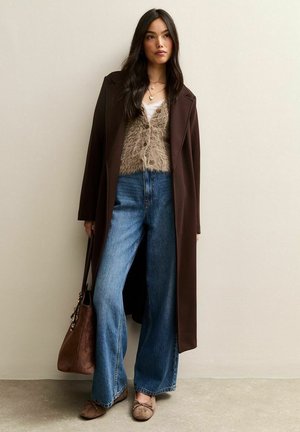 Een bruin lange jas over een fuzzy beige cropped cardigan en hoge taille wijd uitlopende blauwe jeans. Een bruine tas en schoenen maken de look compleet.