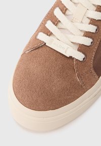 Sneaker in suede marrone con superficie texturizzata, lacci crema e suola in gomma. Presenta un'etichetta logo discreta sul lato.