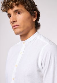 Camicia bianca di lino con colletto alla mandarina, caratterizzata da una placca frontale con bottoni chiari e una finitura del tessuto liscia e texturizzata.