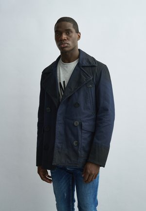 Junger Mann trägt einen marineblauen zweireihigen Mantel, graues Hemd mit schwarzem Text und blaue Jeans, steht vor einem hellgrauen Hintergrund.