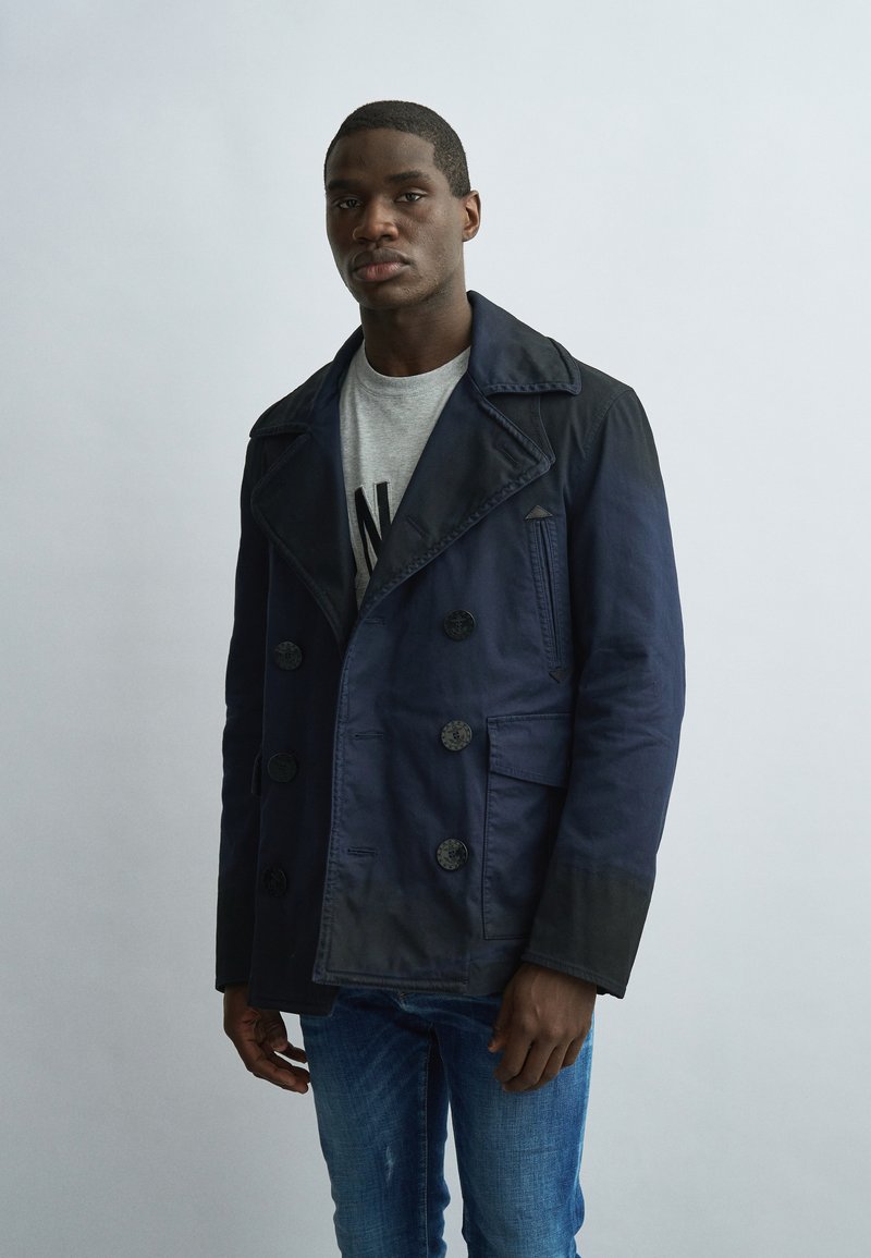 Jeune homme portant un manteau croisé bleu marine, une chemise grise avec un texte noir, et un jean bleu, debout devant un fond gris clair.