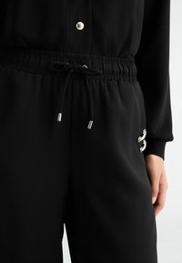 Czarne spodnie z elastycznym pasem i sznurkiem do wiązania, noszone z czarnym topem z złotym guzikiem i ozdobnymi bocznymi akcentami.