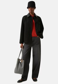 Schwarze Jacke aus Wollmischung mit grauem Kragen, rotem Pullover, dunkelgraue Jeans mit weitem Bein, schwarze flache Schuhe und weiche graue Tragetasche.