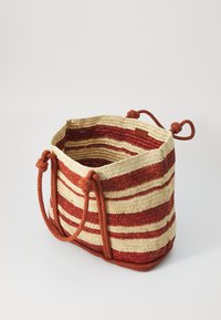 Wiklinowa torba z słomy z poziomymi czerwonymi i kremowymi paskami, z dwoma brązowymi uchwytami z lin i związywaną zapięciem. Wnętrze nie jest podszewkowane.
