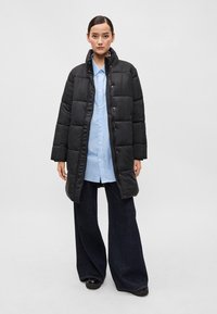 Črna napihnjena jakna s quiltnim vzorcem, visokim ovratnikom in gumbi na pritisk, nosi se preko svetlo modre črtaste srajce in širokih temno modrih kavbojkah.