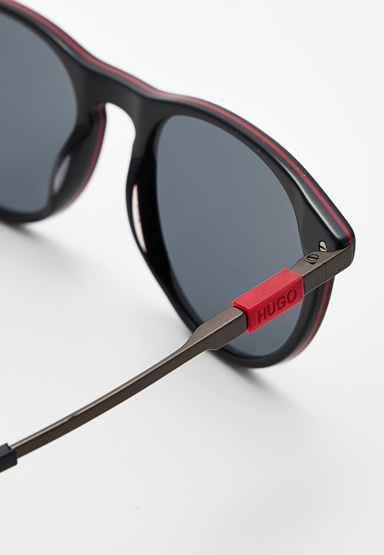 Occhiali da sole neri con montatura tonda, lenti nere e un accento in gomma rossa sulla tempia. Bracci metallici sottili con finitura opaca.