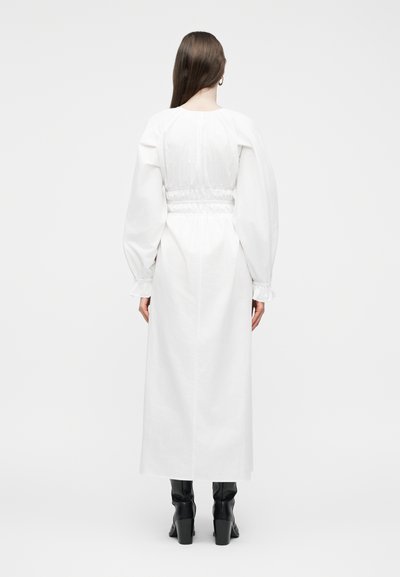 Γυναίκα με μακριά καστανά μαλλιά, φοράει λευκό μίντι φόρεμα με μακριά μανίκια και σφηκοφωλιά στη μέση, στέκεται πλάτη, συνδυασμένο με μαύρα μπότες με τακούνι.