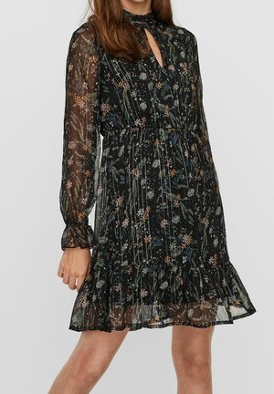 Femme portant une robe longue à manches longues en voile noir à motifs floraux, avec un col montant, un détail goutte d'eau, une taille cintrée et un ourlet à volants.