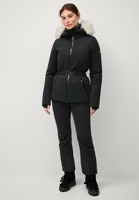 Sort termisk jakke med hætte i faux fur, indsnævret talje, lynlås lukning og sidelommer, parret med matchende sorte bukser og støvler.