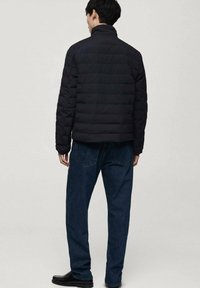 Veste matelassée noire avec col montant, arborant un motif de quilting horizontal, portée sur un jean en denim bleu. Design minimaliste, sans logos visibles.