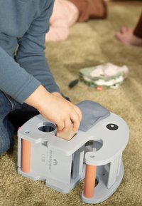 Grijs houten speelgoed met zachte blauwe stofaccenten, met een afgeronde vorm, uitsnijdingen en gekleurde cilindervormige pennen. De hand van een kind speelt ermee.