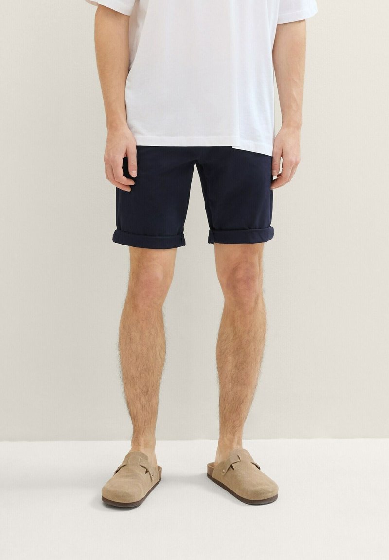 Shorts bleu marine avec ourlets roulés, fabriqués en tissu léger. Portés avec des chaussures beige à enfiler. Chemise blanche unie surdimensionnée.